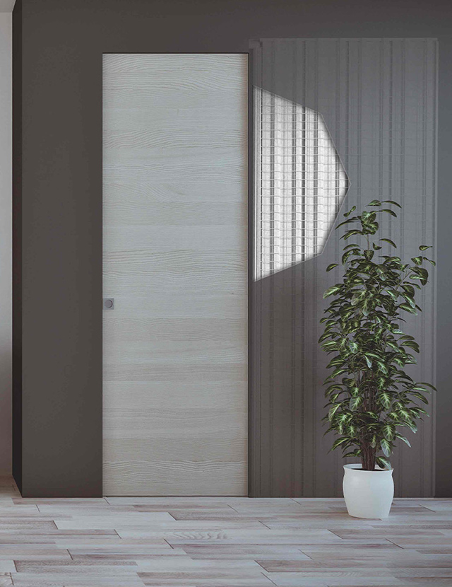 Dettaglio di una porta scorrevole a scrigno con finitura in legno chiaro venato. Una sezione visiva sulla parete grigia mostra la robusta struttura in lamiera del controtelaio interno.