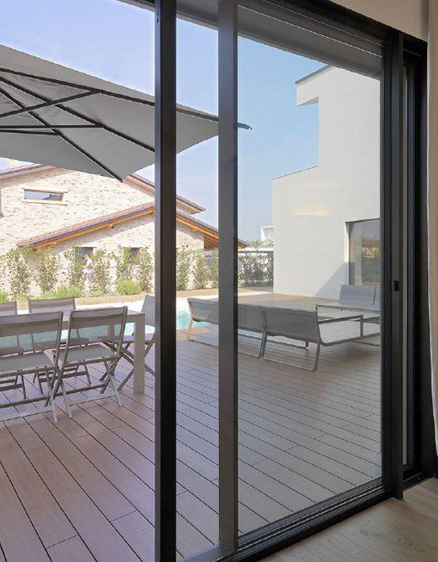 Dettaglio del profilo minimale di un infisso scorrevole Schüco Living Slide scuro, aperto verso un elegante patio esterno in legno con piscina.