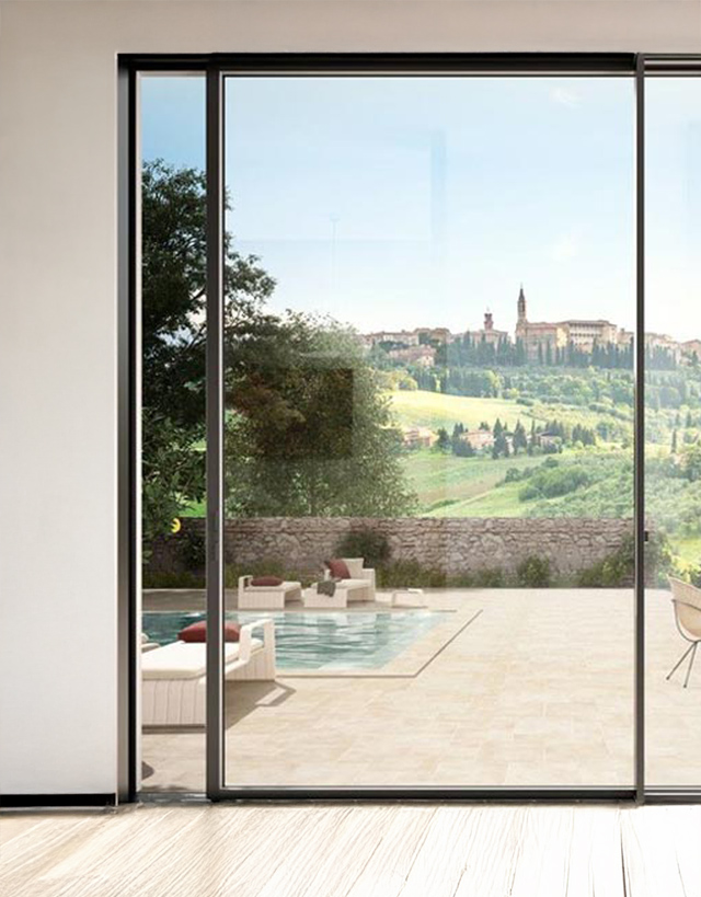 Grande vetrata scorrevole panoramica Schüco con profili in alluminio sottili, aperta su un patio con piscina e un suggestivo borgo collinare.