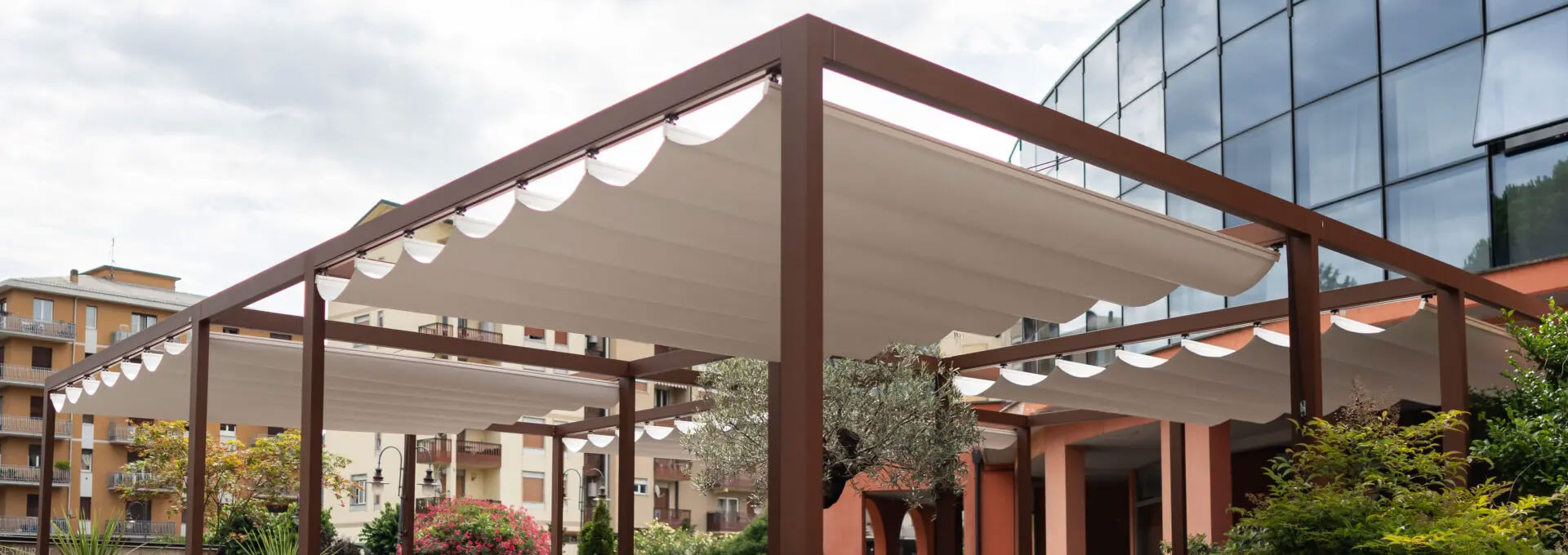 Pergotende KE a Roma: coperture versatili per grandi spazi all'aperto Pergotenda KE con robusta struttura in alluminio marrone e telo retrattile a falde, installata per ombreggiare un ampio cortile esterno.