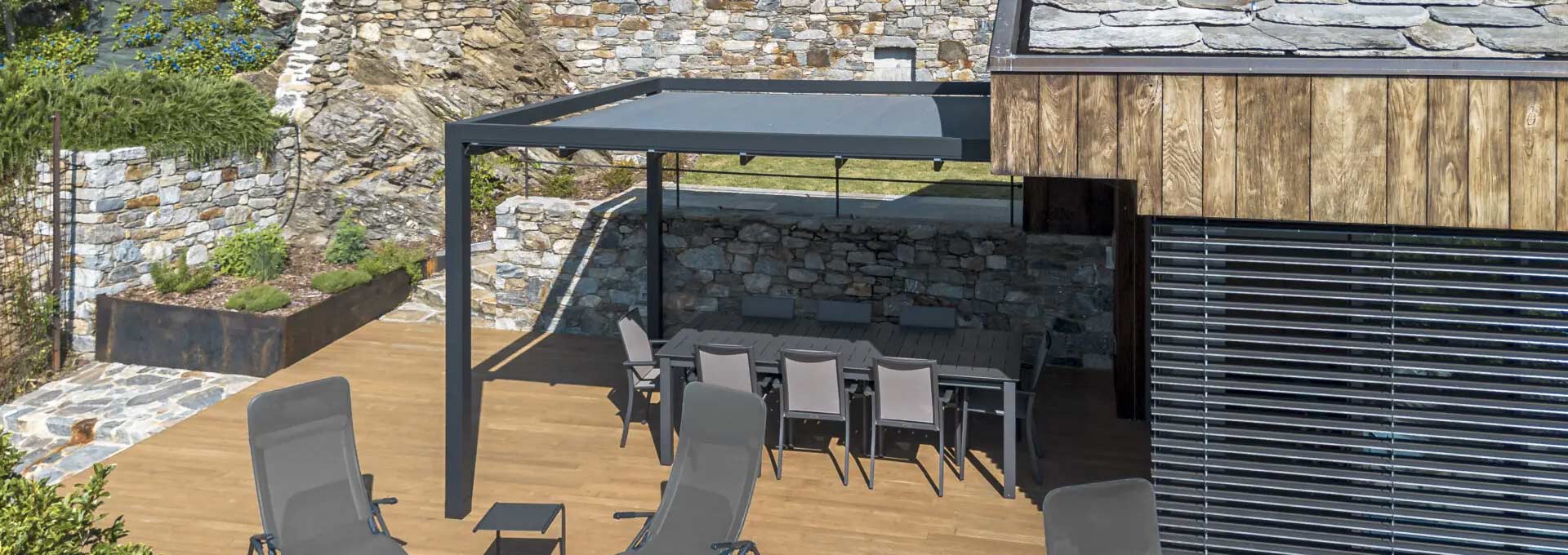 Pergole addossate KE: crea la tua zona pranzo outdoor Pergola addossata KE color grigio antracite che protegge un tavolo da pranzo esterno, perfettamente integrata nell'architettura di una casa in pietra e legno.