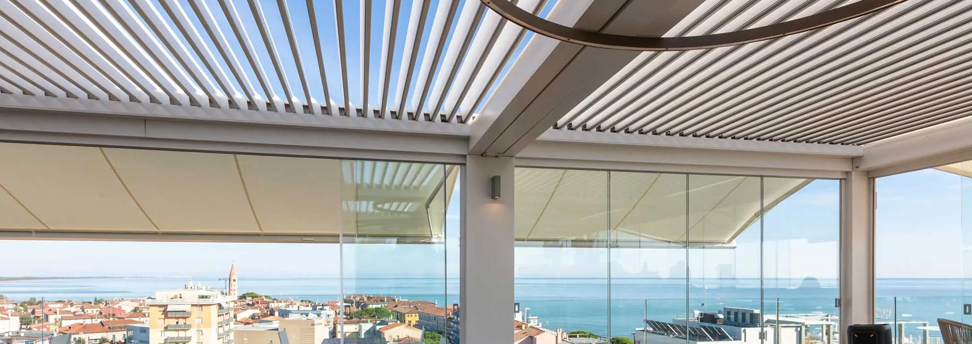 Vivere il terrazzo tutto l'anno: pergole bioclimatiche KE con vetrate panoramiche Terrazzo panoramico coperto da una pergola bioclimatica KE bianca con lame orientabili e chiusure perimetrali in vetro scorrevole, con vista sul mare.