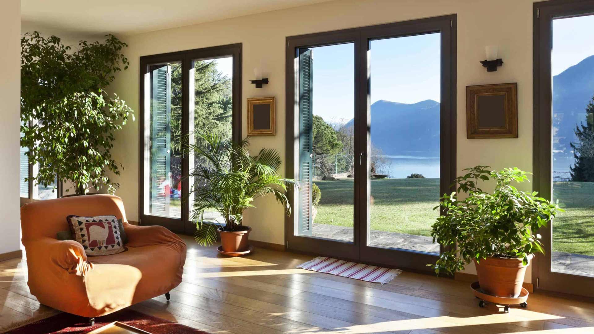 Luminoso soggiorno con divano arancione e grandi porte finestre affacciate su un panorama vista lago, impreziosite da eleganti persiane a cardine verdi aperte verso l'esterno.