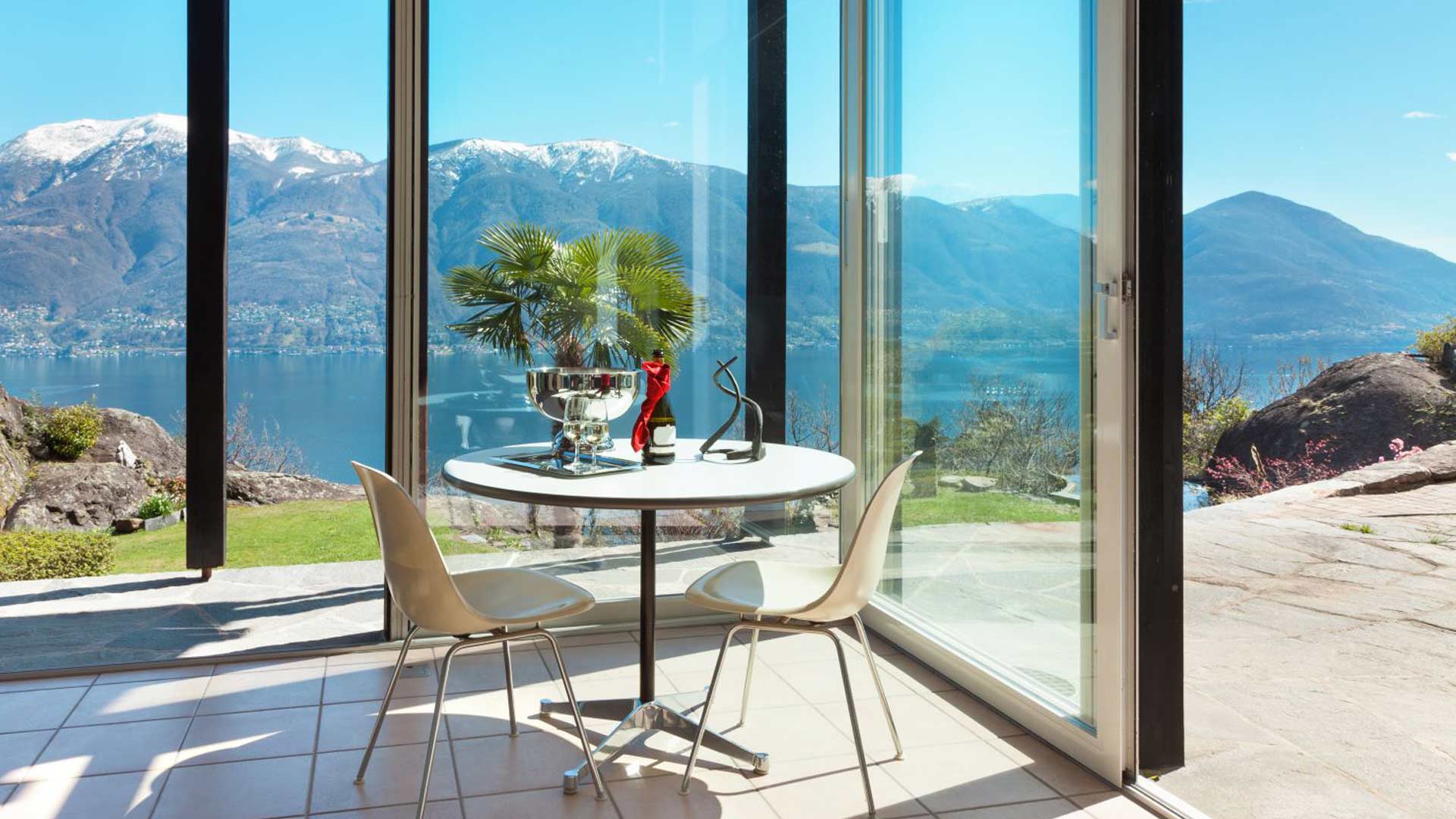 Luminosa area relax con tavolino, sedie e vista mozzafiato su lago e montagne, protetta da eleganti vetrate panoramiche scorrevoli totalmente trasparenti e prive di profili verticali.