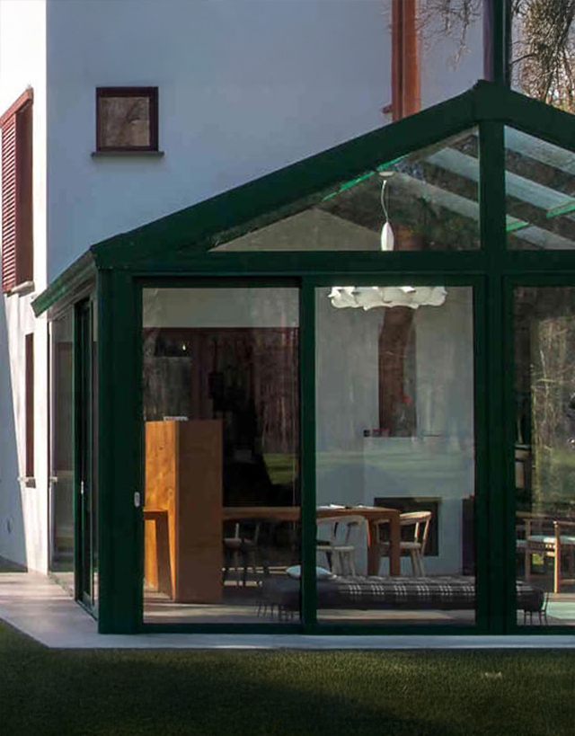 Elegante giardino d'inverno addossato a una villa moderna, realizzato con struttura portante scura e ampie vetrate panoramiche che proteggono un'accogliente zona pranzo interna.