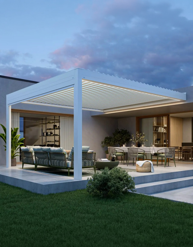 Elegante pergola bioclimatica bianca addossata alla parete di casa al tramonto. Le lame orientabili del tetto sono chiuse e l'illuminazione a LED perimetrale accesa illumina la zona pranzo e relax sottostante.