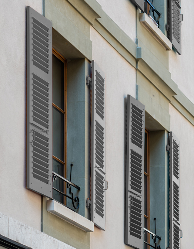 Facciata di un palazzo con alte portefinestre protette da persiane in alluminio a cardine di colore grigio, aperte per far entrare la luce.