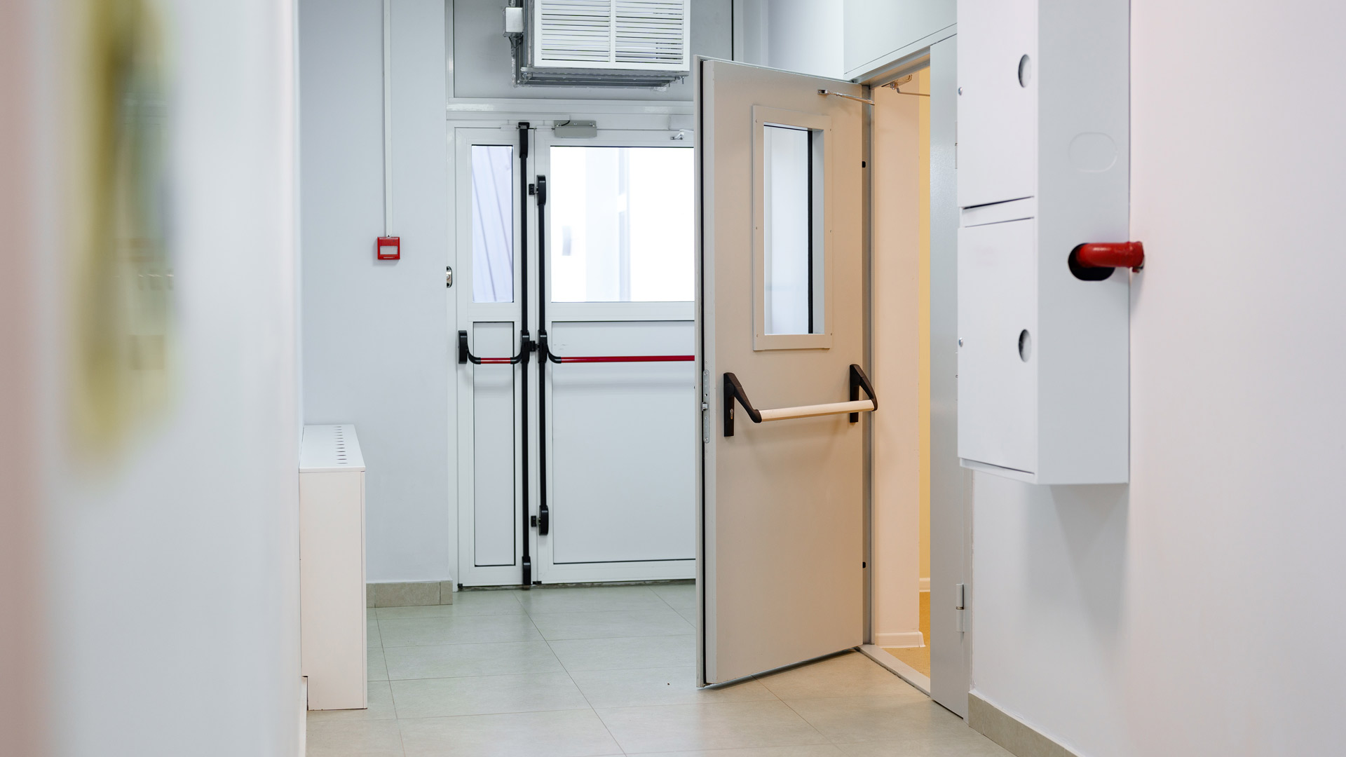 Corridoio interno di un edificio con una porta antincendio tagliafuoco aperta, dotata di maniglione antipanico rosso e piccolo oblò in vetro di sicurezza.