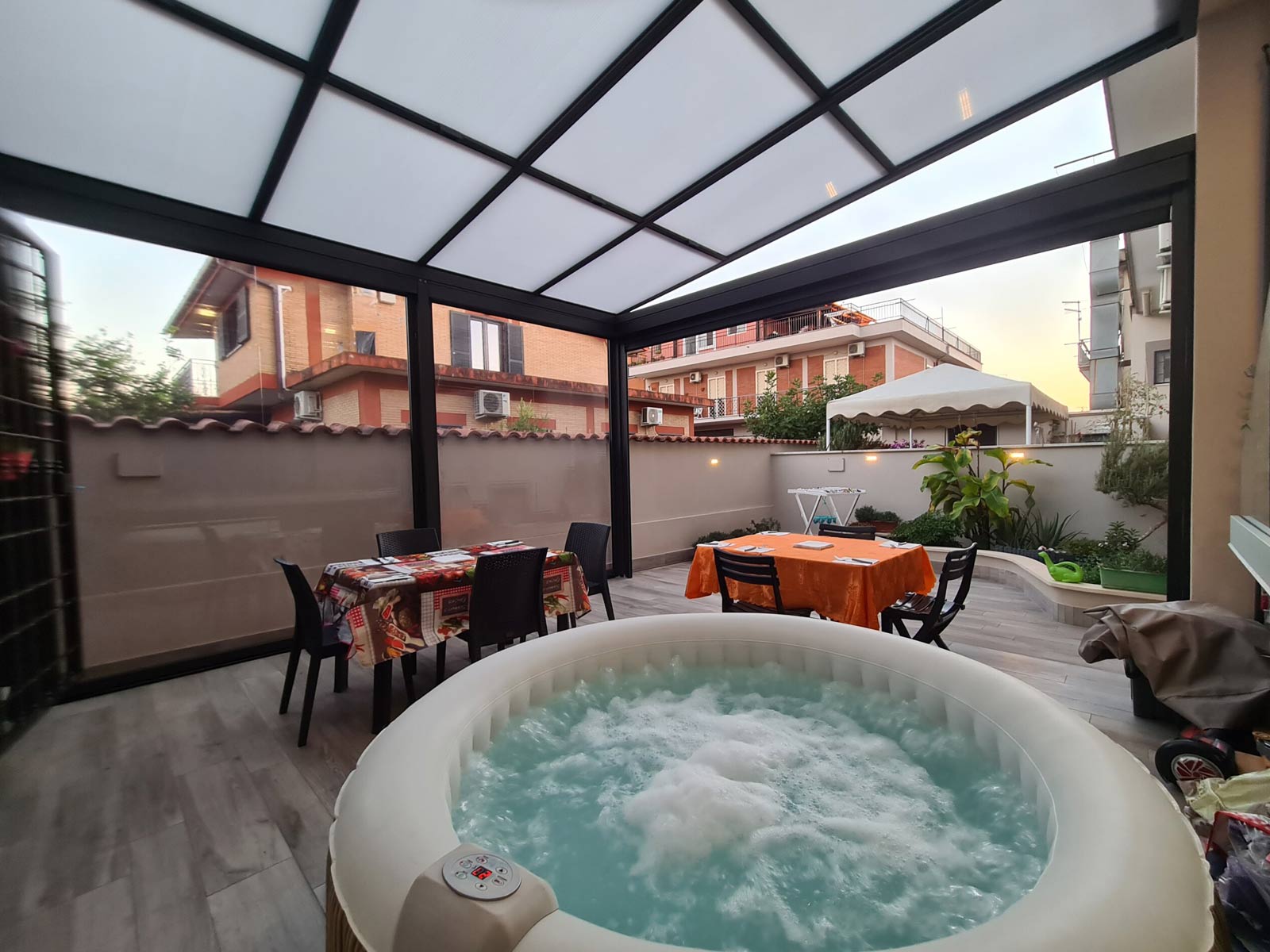 Pergola bioclimatica scura chiusa con vetrate scorrevoli, arredata con tavolo da pranzo e vasca idromassaggio