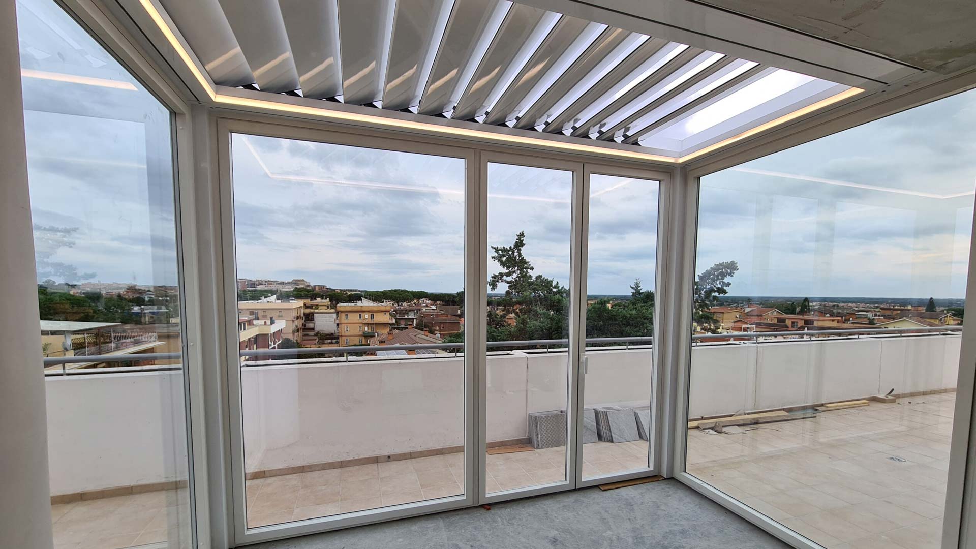 Interno di una pergola bioclimatica bianca con lame frangisole e vetrate scorrevoli su un attico a Roma