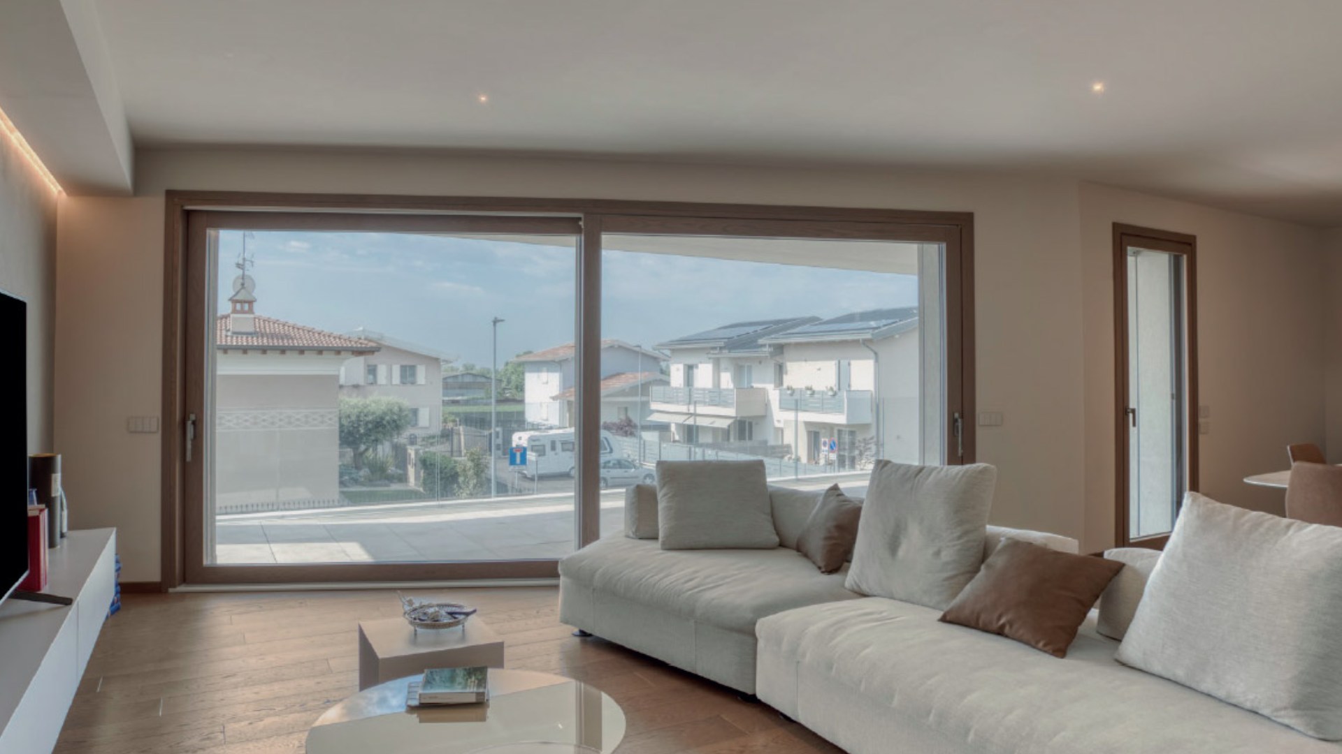 Luminoso soggiorno moderno con un grande infisso scorrevole in legno-alluminio DMR finitura rovere, affacciato su un tranquillo paesaggio residenziale.