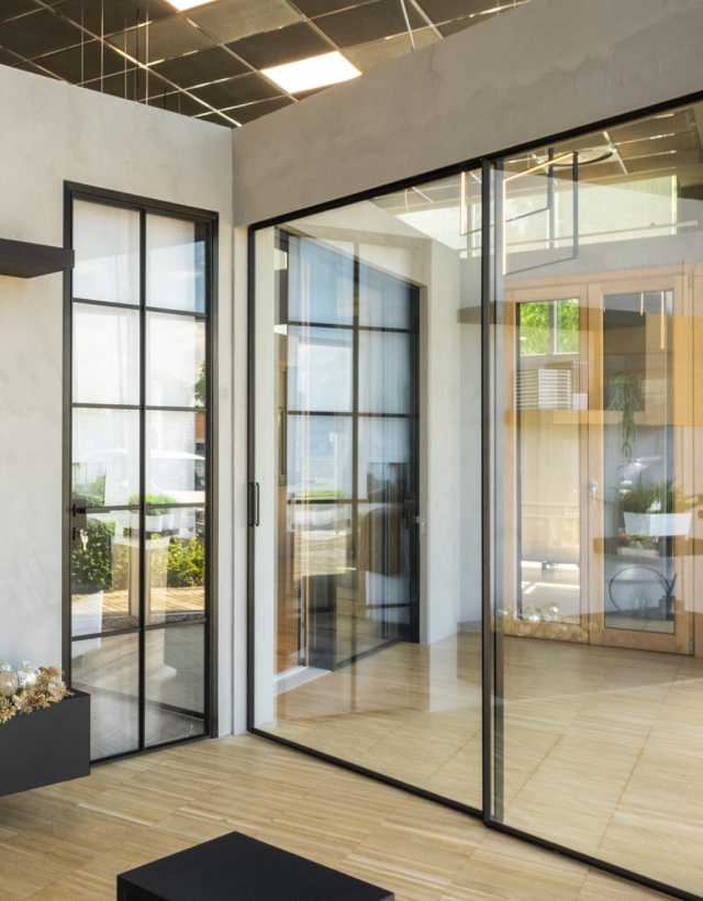 Allestimento in showroom con eleganti infissi a profilo minimale nero, che mostra un'ampia vetrata scorrevole affiancata da una porta a battente con inglesine in stile industriale.
