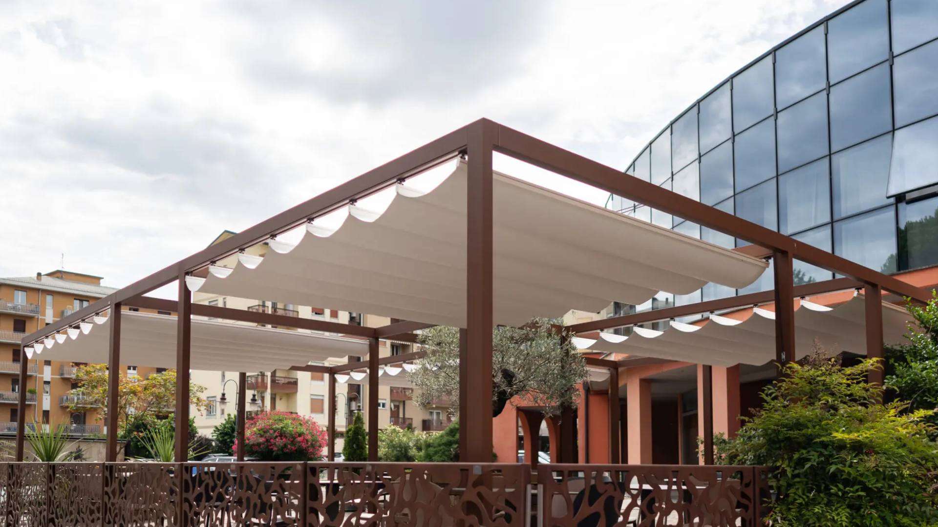 Grande pergola in alluminio marrone KE con tetto scorrevole a impacchettamento in tessuto chiaro, installata in un ampio spazio esterno a Roma.