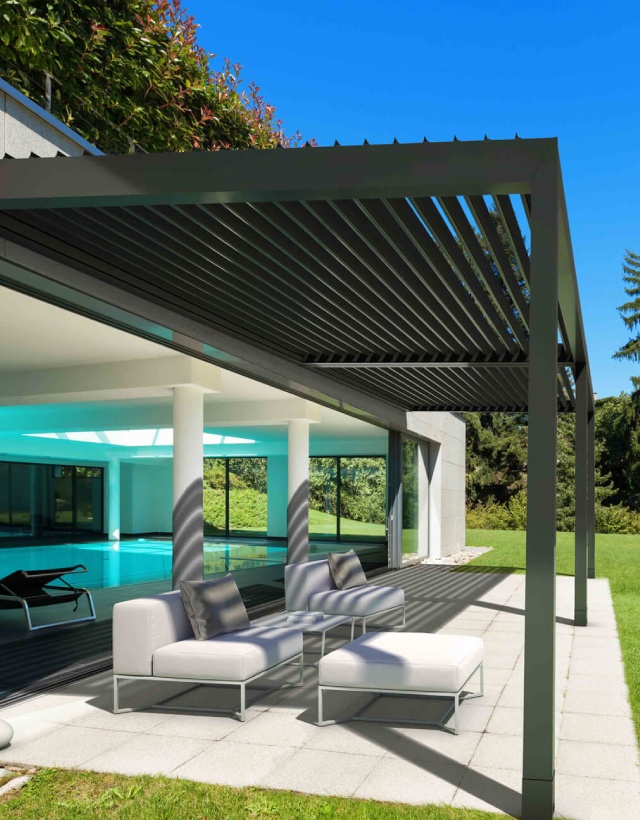 Elegante pergola bioclimatica KE in alluminio grigio scuro con lame frangisole orientabili, addossata a una villa moderna con piscina interna.