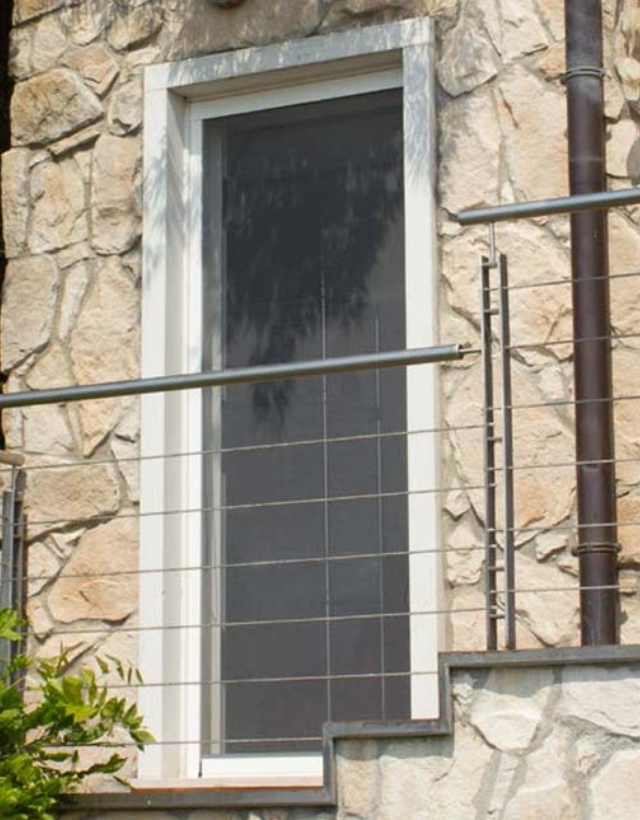 Dettaglio di una finestra verticale installata su una facciata in pietra grezza, protetta da una zanzariera Bettio a scorrimento verticale perfettamente integrata.